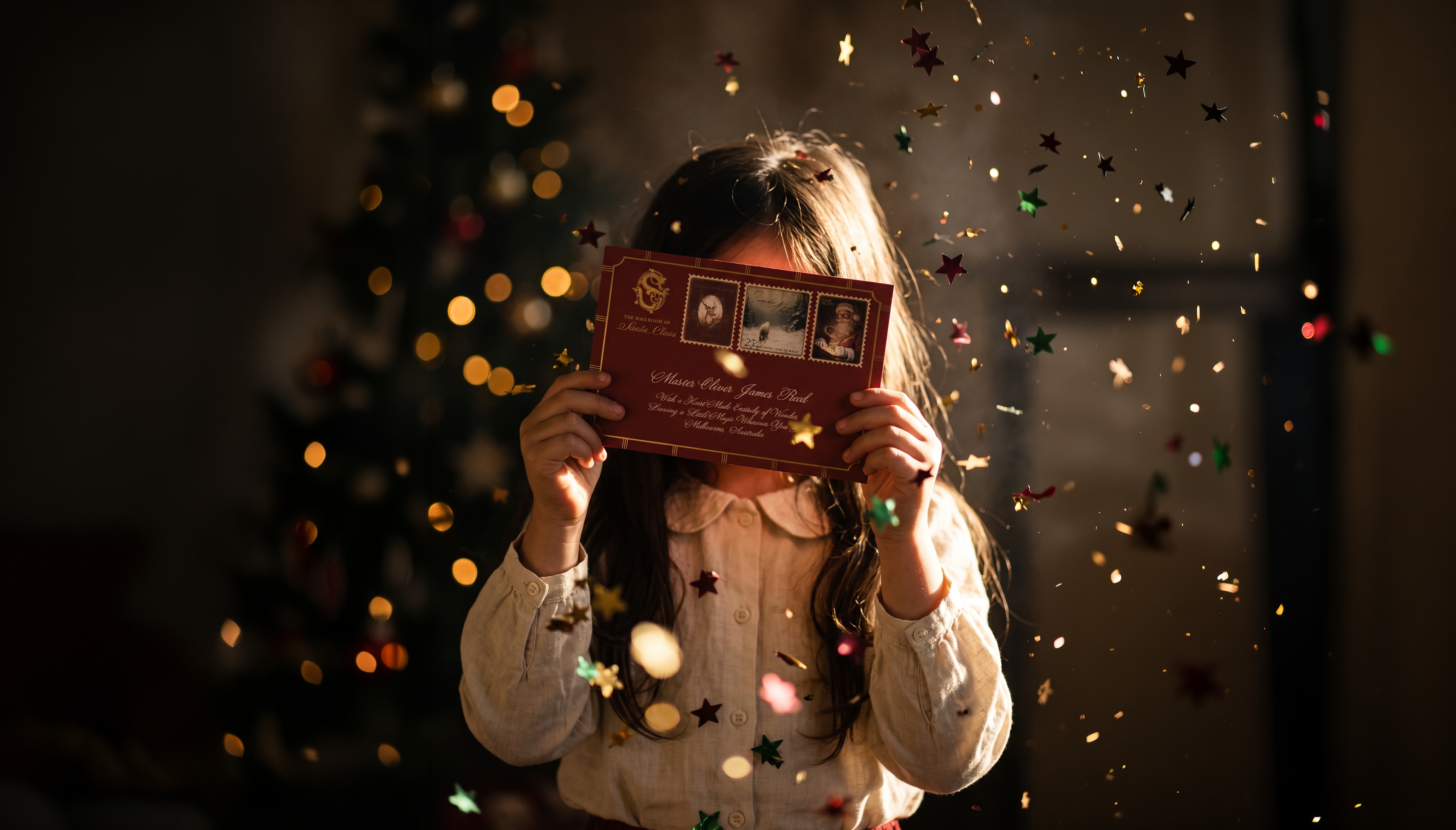 A young girl holding up her personalised letter from Santa Claus in front of a Christmas tree, with gold stars falling around her