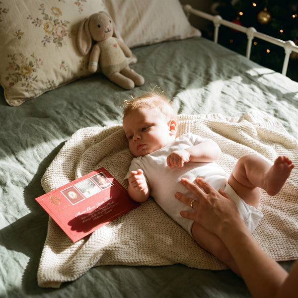 Baby’s First Christmas Letter From Santa Claus - A newborn baby resting on a knitted blanket beside a Baby's First Christmas letter from Santa in a dark red envelope