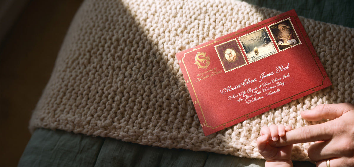 A baby's hand gripping a parent's finger beside a Baby's First Christmas letter from Santa in a dark red envelope