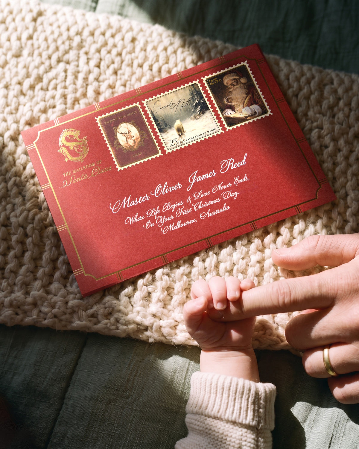 A baby's hand gripping a parent's finger beside a Baby's First Christmas letter from Santa in a dark red envelope