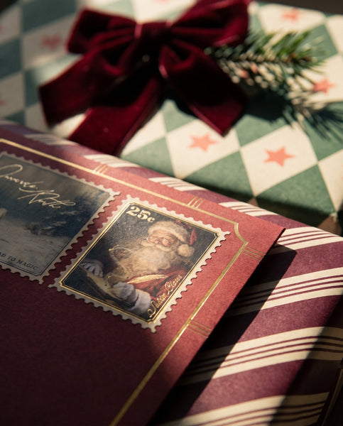 Personalised Letter From Santa Claus - Close up of a dark red Santa letter envelope with a gold framed Santa Claus stamp, resting beside a wrapped gift and a burgundy velvet bow