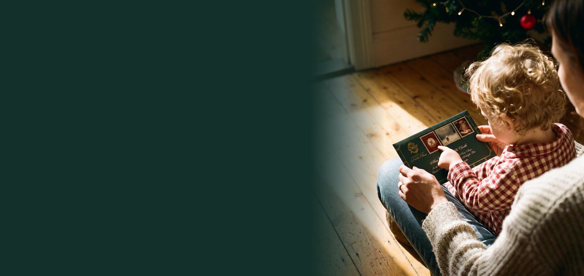A toddler on mum's lap by the Christmas tree holding a green Santa letter envelope